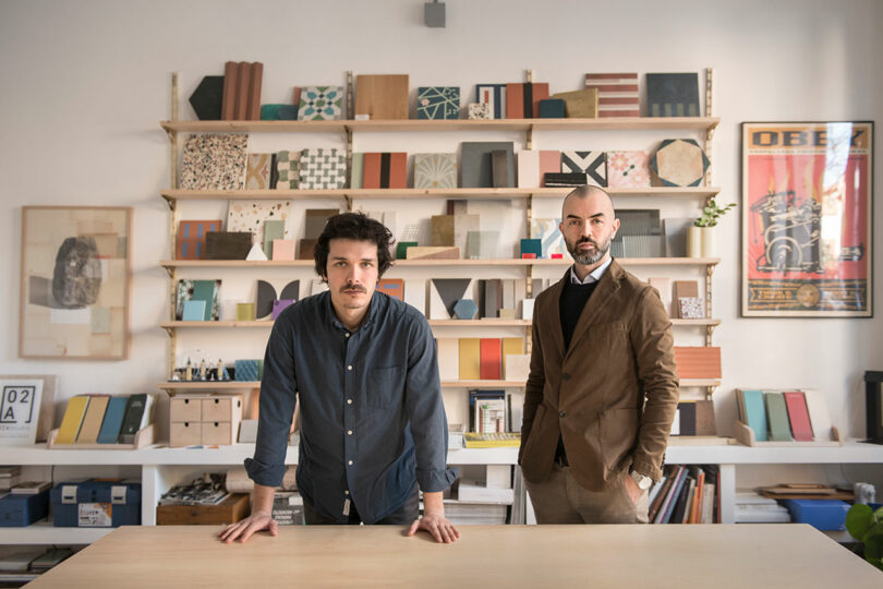 Two men standing over a desk with filled bookshelves behind them.