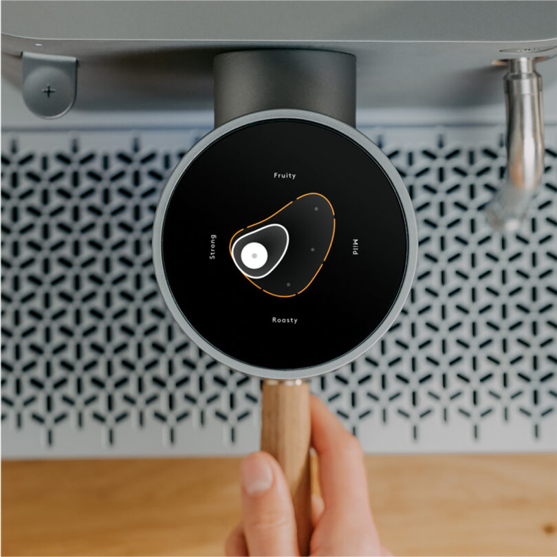 A person holding an AI-enhanced coffee and espresso machine portafilter handle, with the machine's overhead circular color digital display showing the flavor profile of a roast.