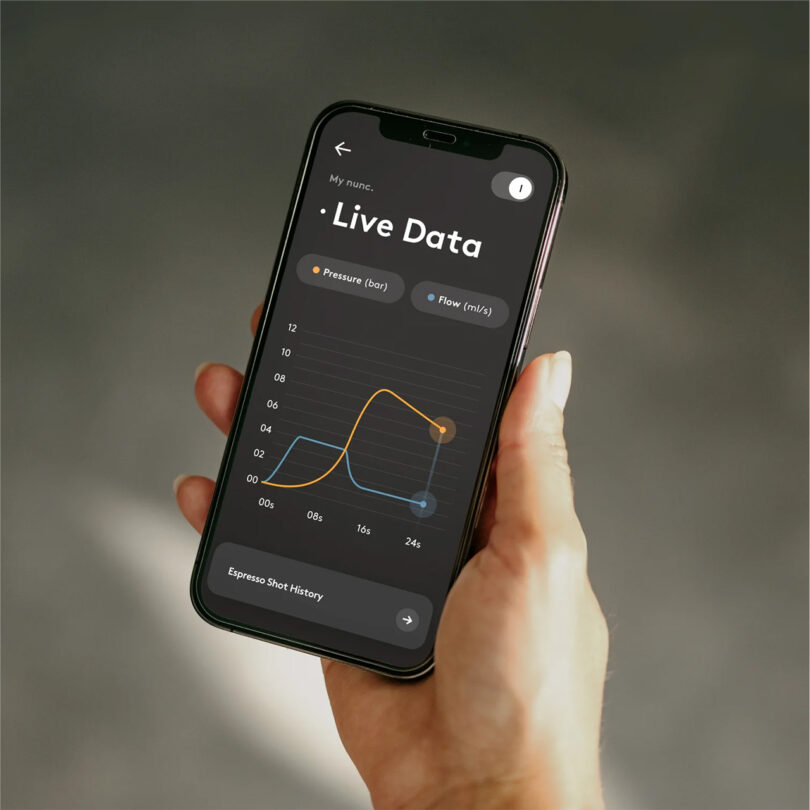A person holding up a smartphone with live data graph showing pressure and flow from an AI-enhanced coffee machine on the screen