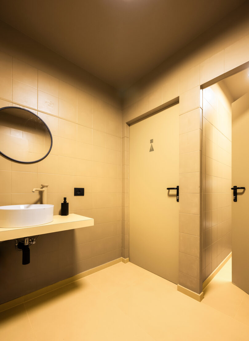 A modern bathroom with beige tiles, featuring a wall-mounted sink, a round mirror, and a NOA glass shower enclosure.