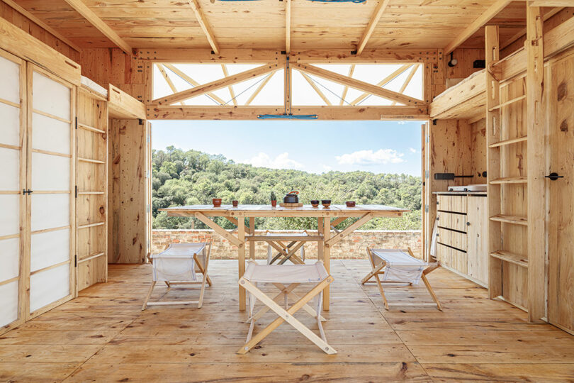 Rustic wooden cabin interior with open double doors leading to a forest view, featuring a simple dining table and chairs.