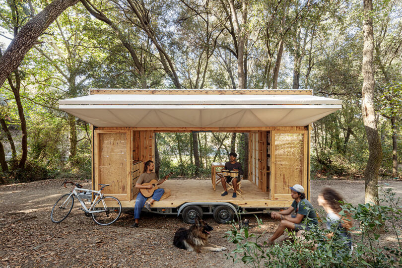 Three individuals and a dog relaxing by a mobile wooden structure in a forest setting.