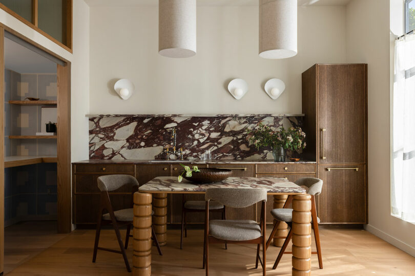 Modern dining room with a marble backsplash, round wooden table, and unique cylindrical chairs, complemented by pendant lighting and wooden cabinets.