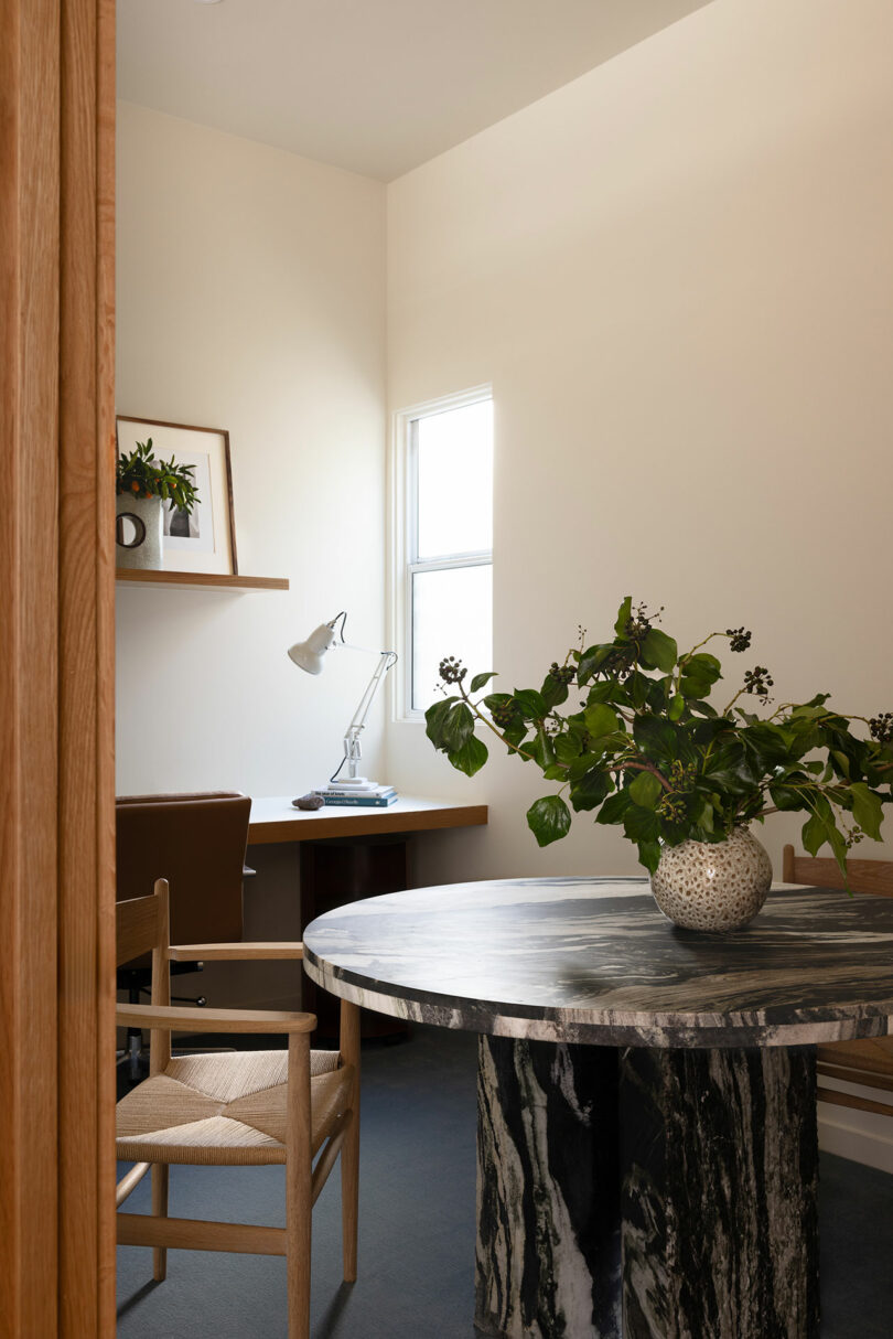 A minimalistic home office with a marble desk, wooden chair, desktop lamp, and plants on shelves by a window.